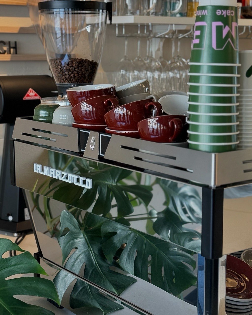A sleek stainless steel espresso machine is adorned with neatly stacked burgundy and white cups and a row of green paper cups, set against a chic café backdrop with hanging glasses and leafy decor, highlighting a modern coffee shop atmosphere.
