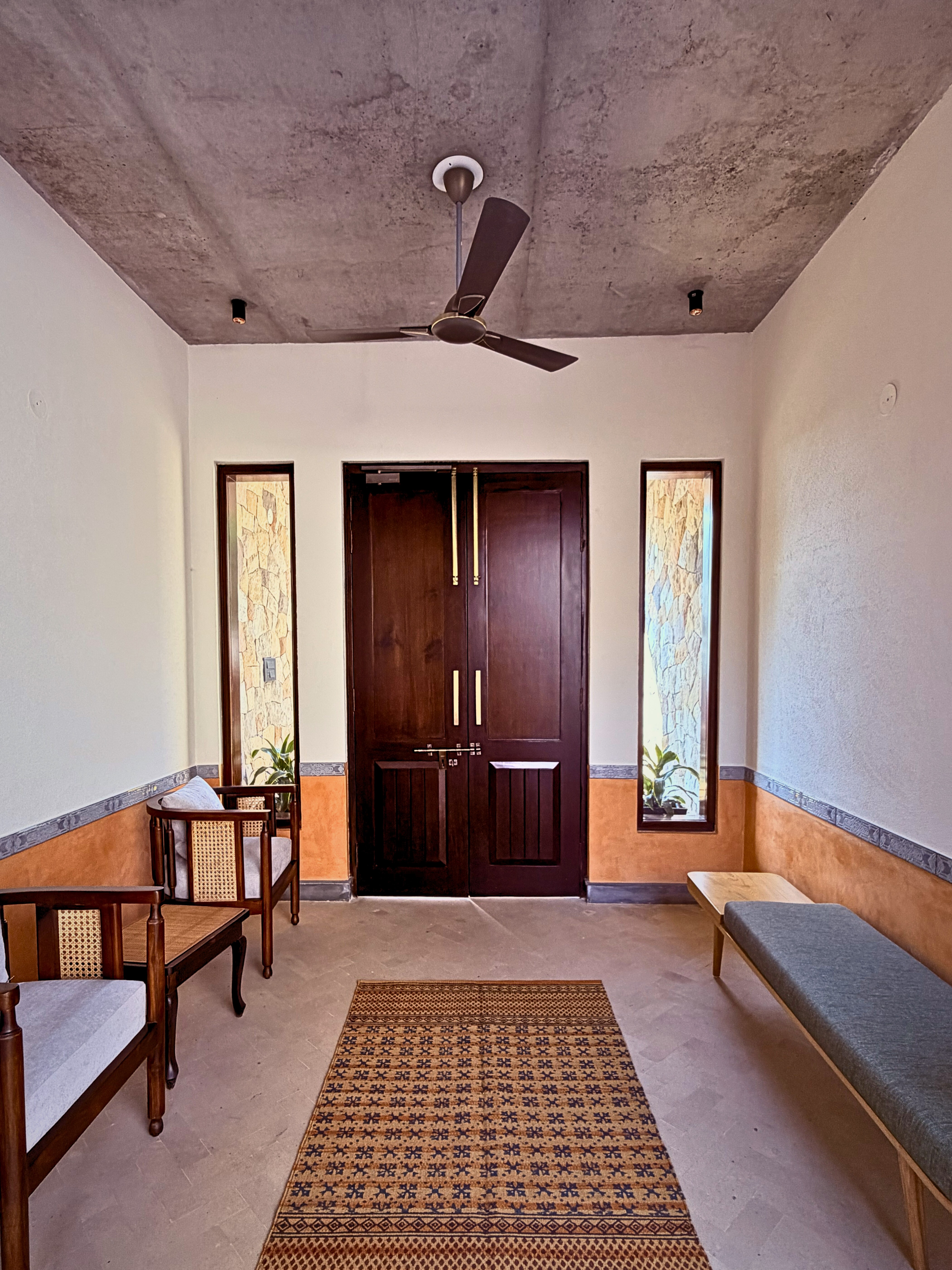 Transitional calm: This foyer blends modern and rustic elements, featuring a simple bench, cane armchairs, and a patterned rug set against a backdrop of textured walls and concrete.