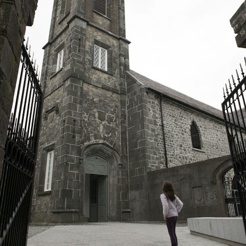 一个人站在一座历史悠久的石砌教堂附近，教堂有一个拱形入口和锻铁大门，他正仰望这座建筑。