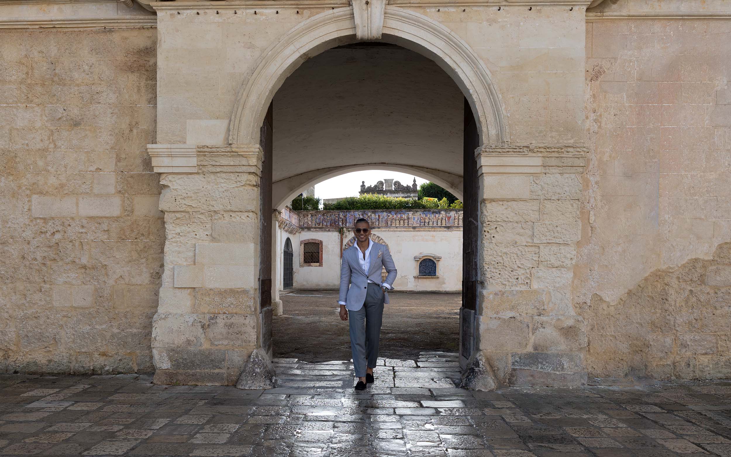 A man wearing a light gray tailored jacket walks through an Italian stone archway, representing Don Marrón’s refined approach to modern Italian menswear and craftsmanship.