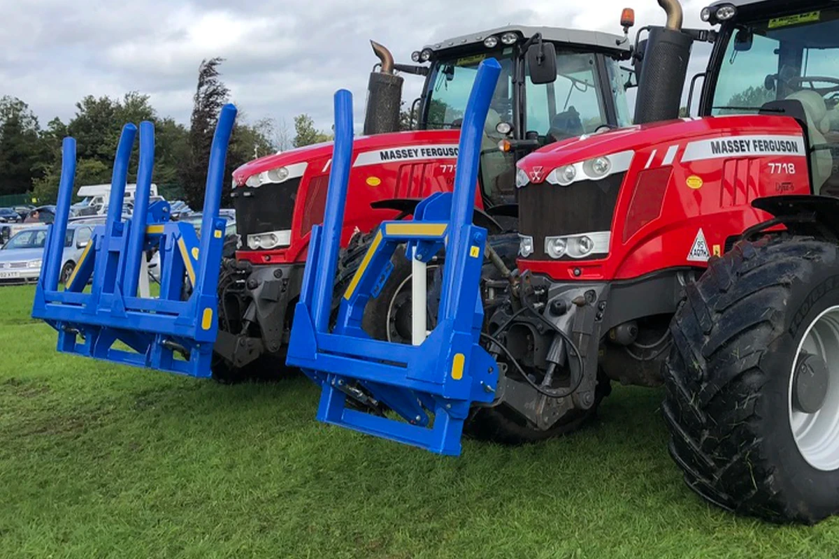 Two Massey Ferguson red tractors in a field side by side with a blue Hydraulic folding bale handlers attached