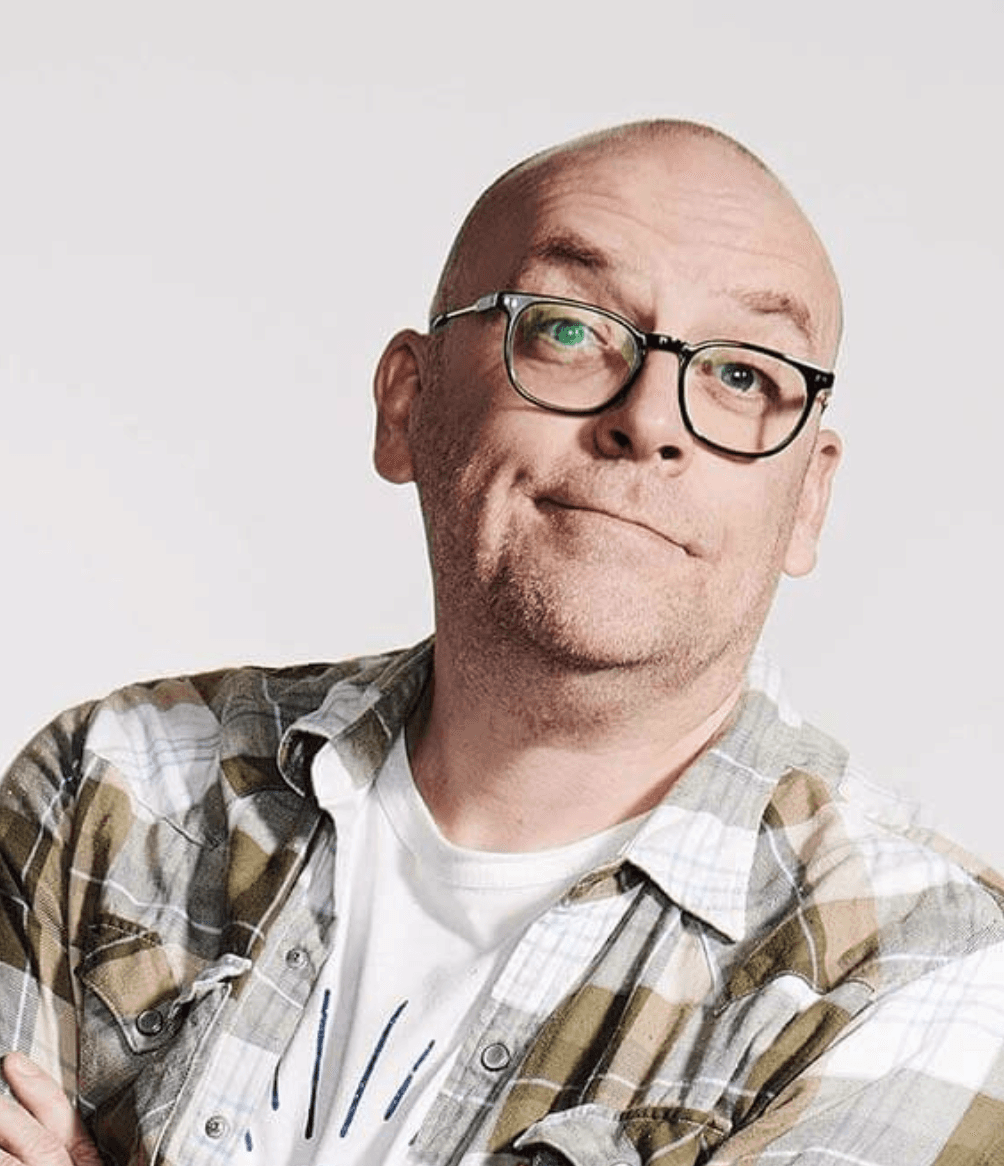 A portrait of Phil Booth - a smiling person wearing glasses and a patterned shirt poses against a light background.