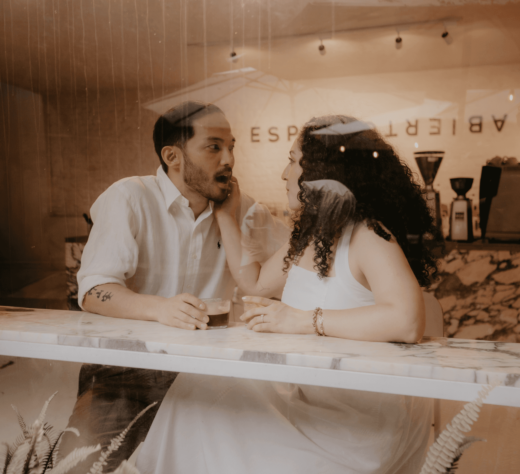 A couple stands on a sandy hill at sunset, facing away, with arms raised in a relaxed "hang loose" gesture. The scene conveys tranquility and joy.