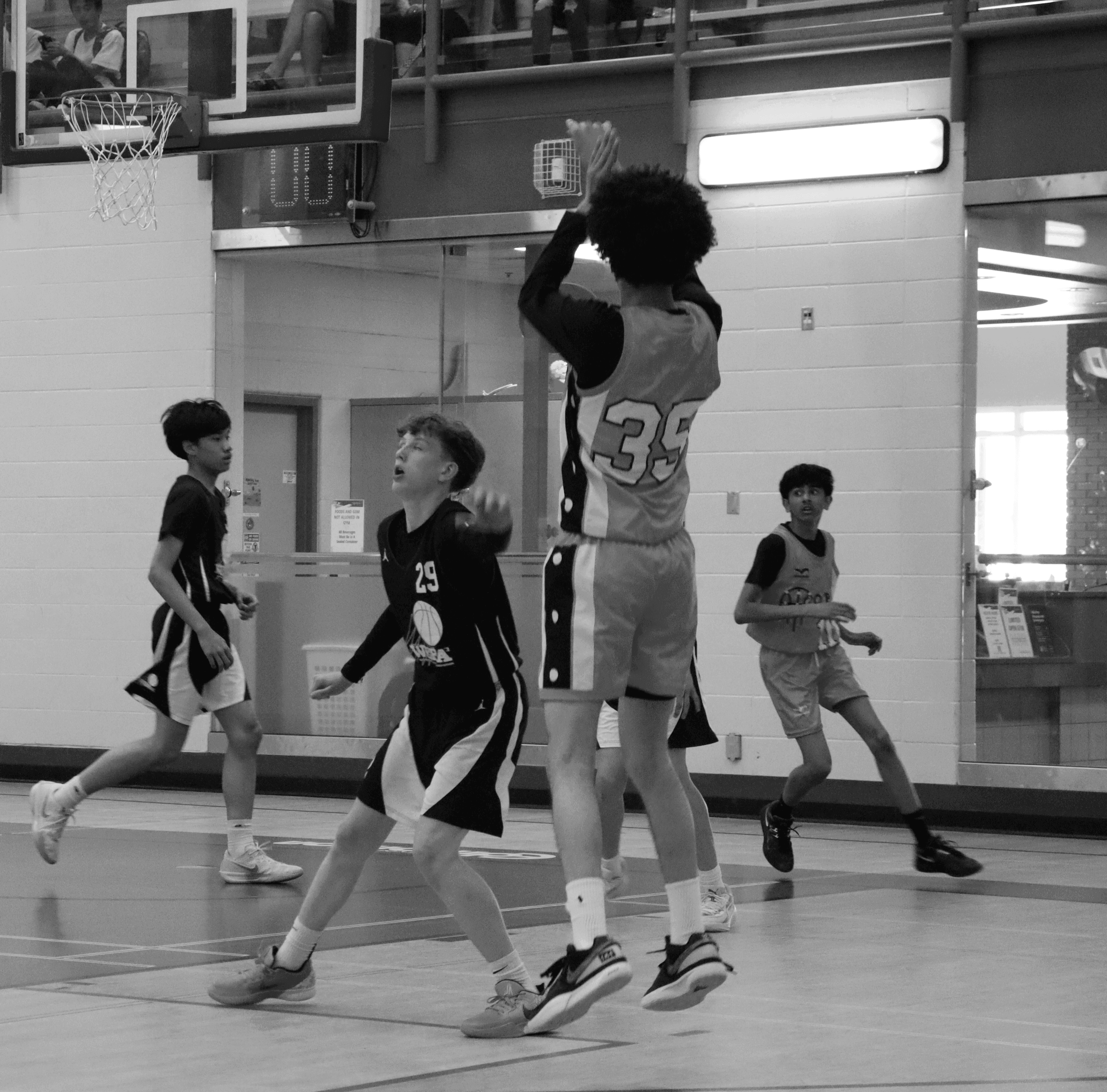 A youth basketball player in an orange jersey dribbling aggressively past a defender during a game, with a referee and spectators visible in the background on an indoor court.