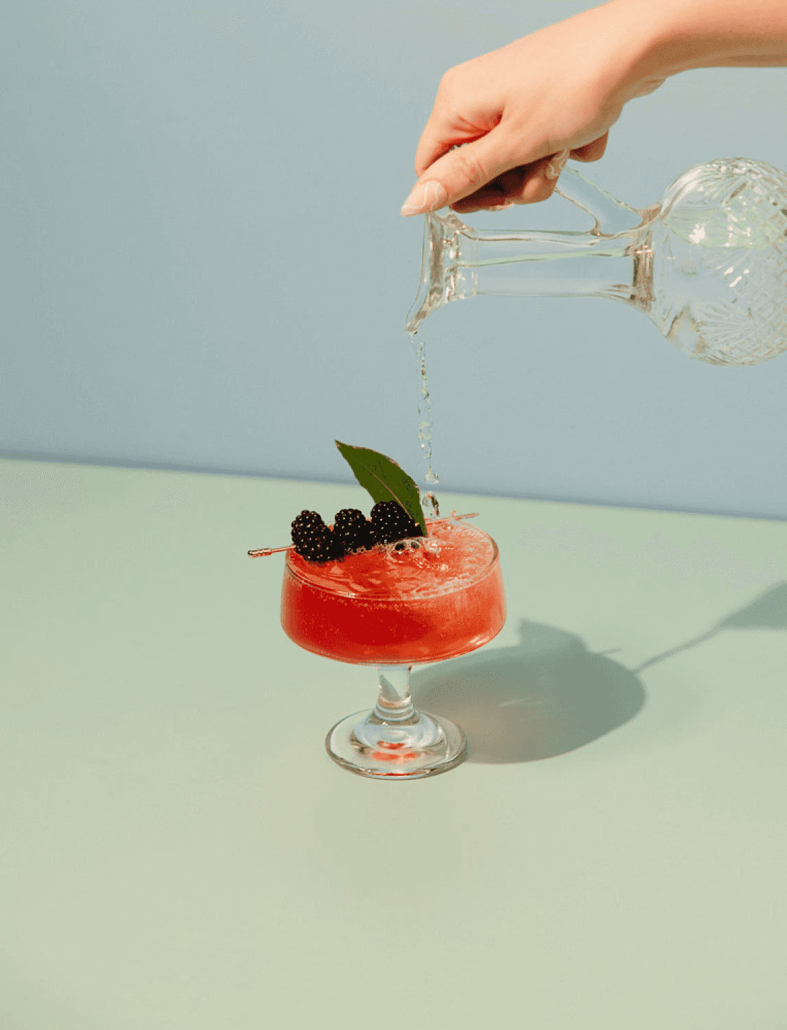 A hand from above pouring seltzer into a bright red drink garnished with blackberries