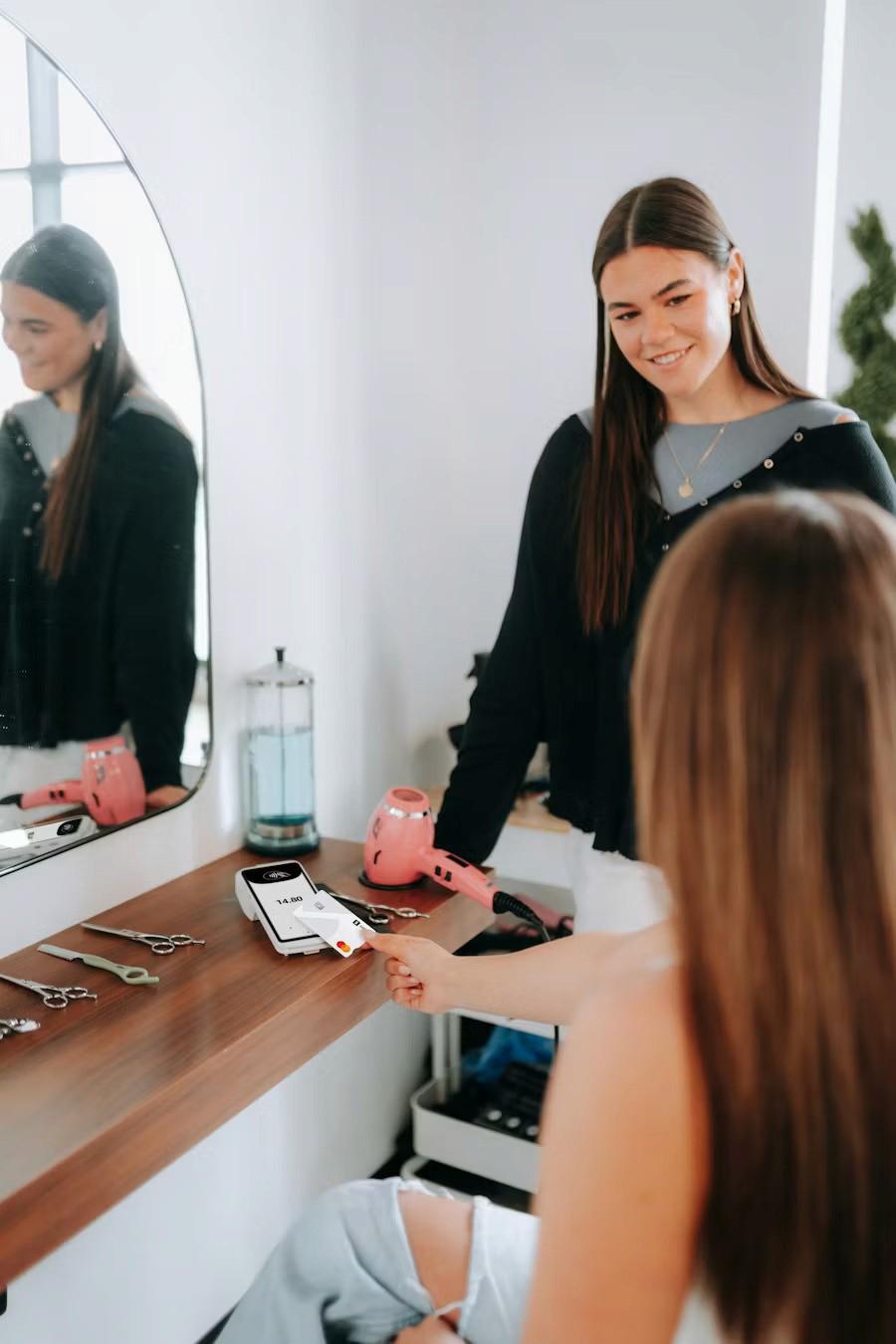 Two women are in a hair salon; one sitting and the other standing by a wooden vanity with styling tools like scissors and a hairdryer, reflecting in a large wall-mounted mirror.