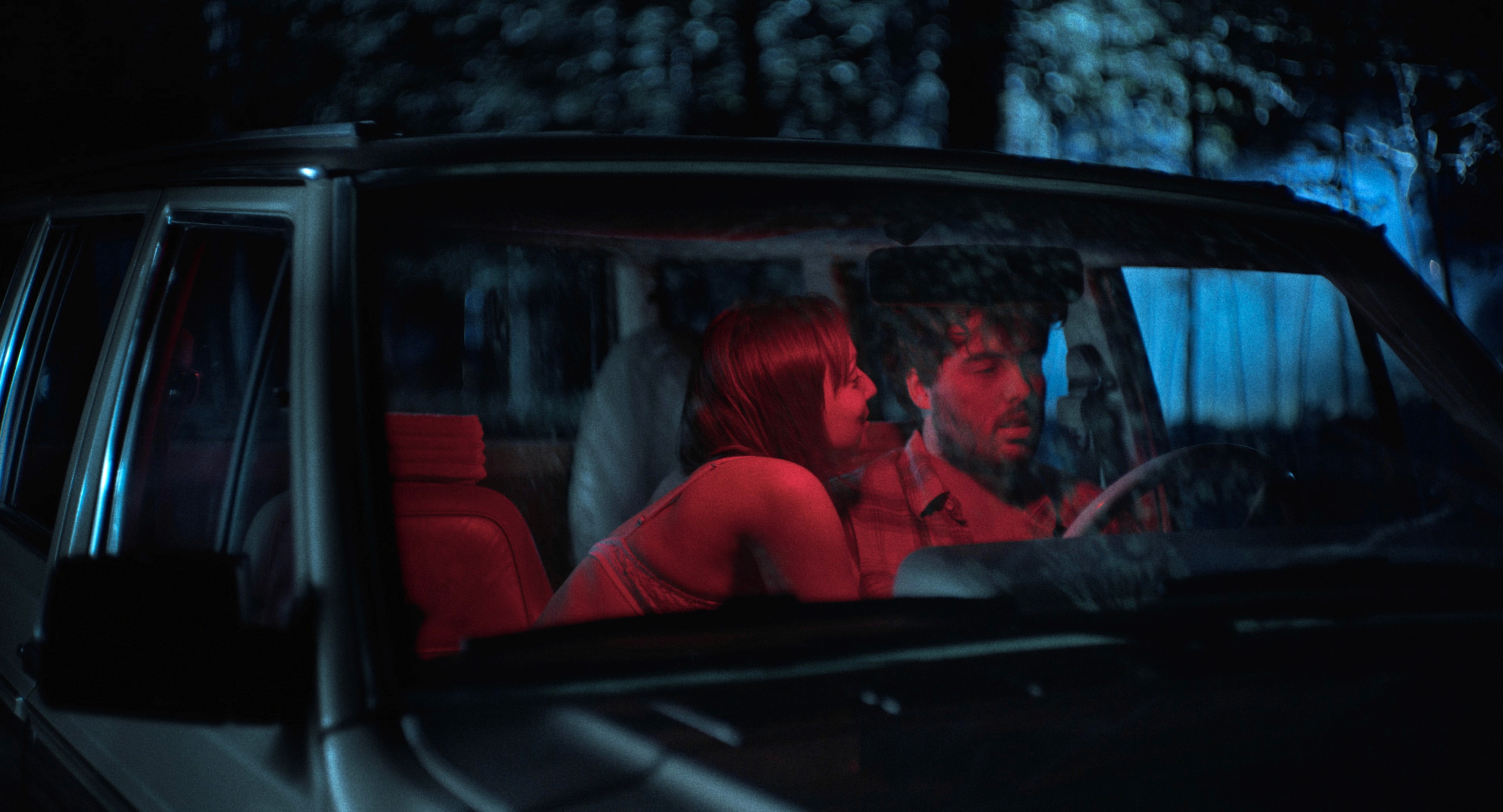 Une femme et un homme assis dans un véhicule stationné la nuit. La femme est penchée vers l'homme. Scène tirée du court métrafe NU