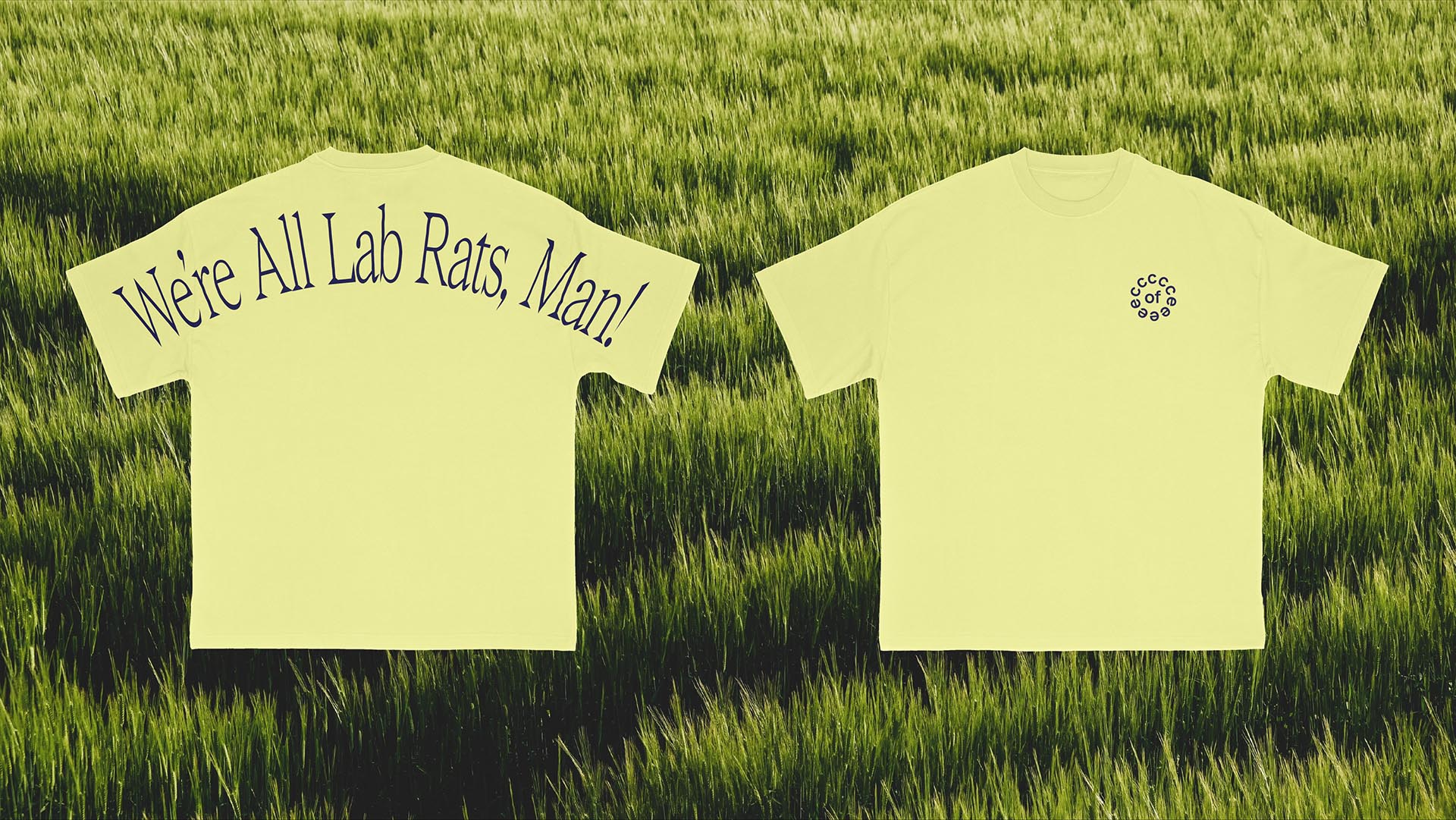 Two pale yellow t-shirts lie flat on bright green grass. The left shirt displays the text "We're All Lab Rats, Man!" in dark blue. The right shirt features a small, dark circular logo near the chest.