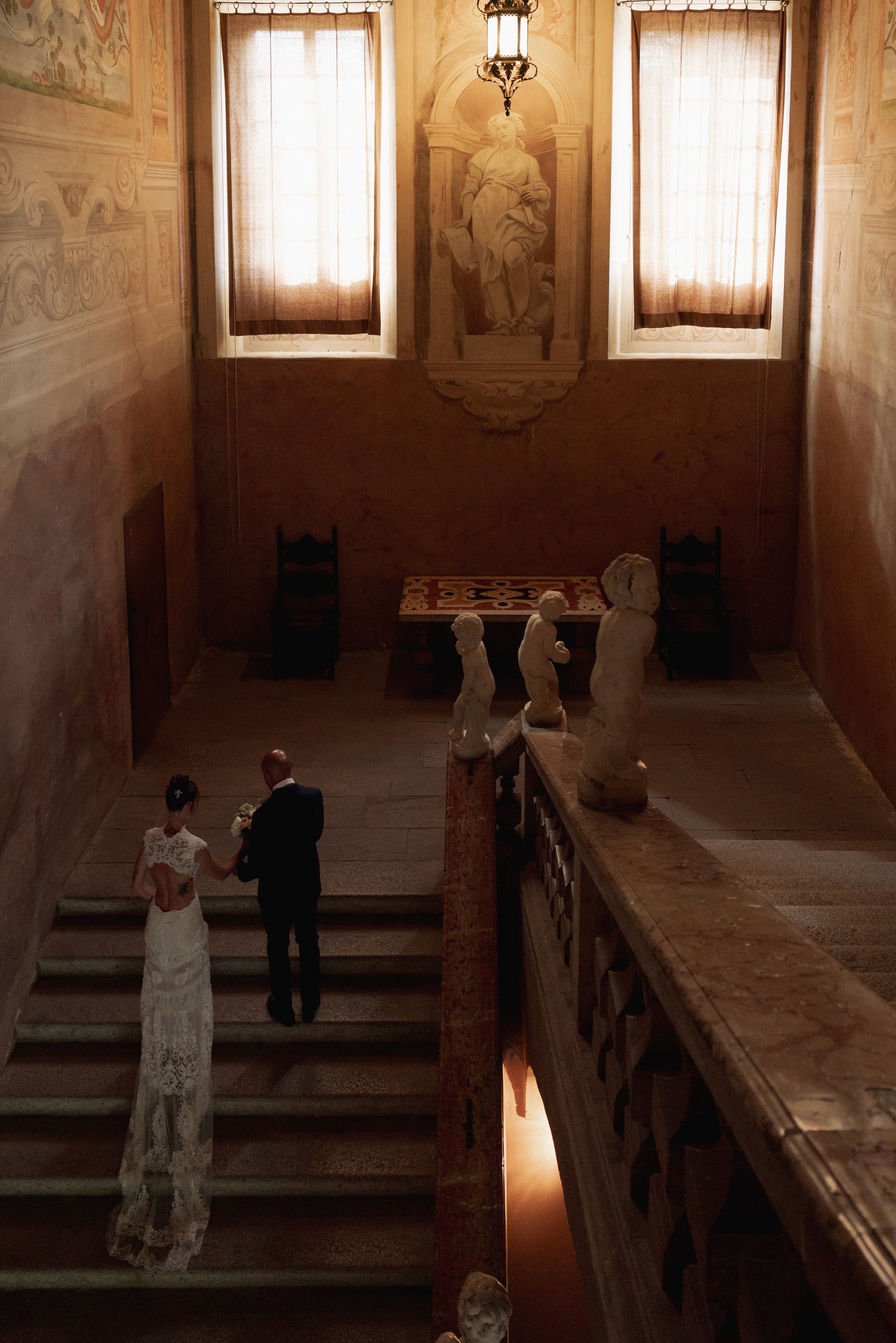Sposi scendono la scala della Rocca di Soragna dopo cerimonia, luce dorata dalle finestre illumina il reportage matrimoniale