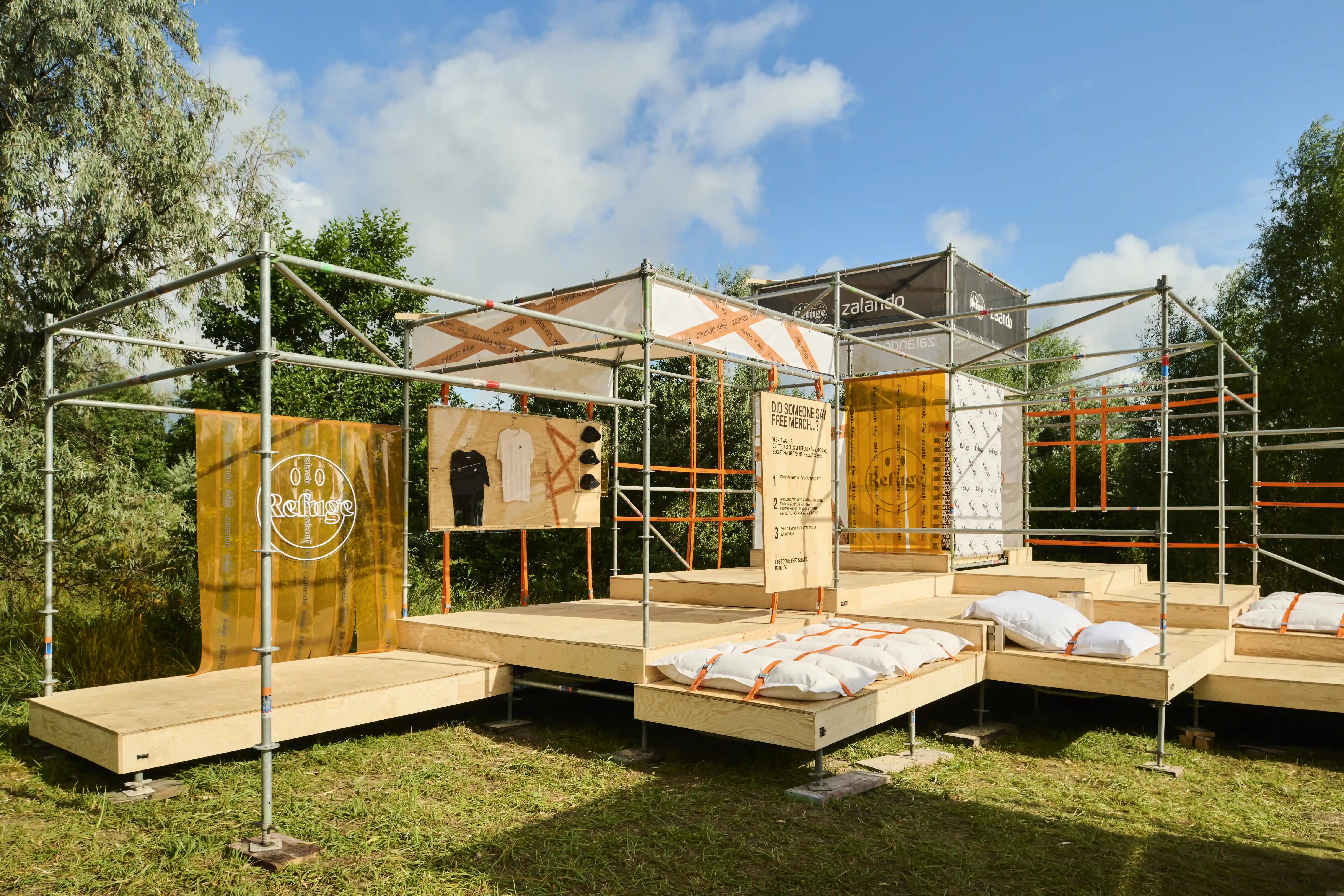 Open-air lounge of scaffolding and plywood decks with pillow beds and Refuge logo wall.