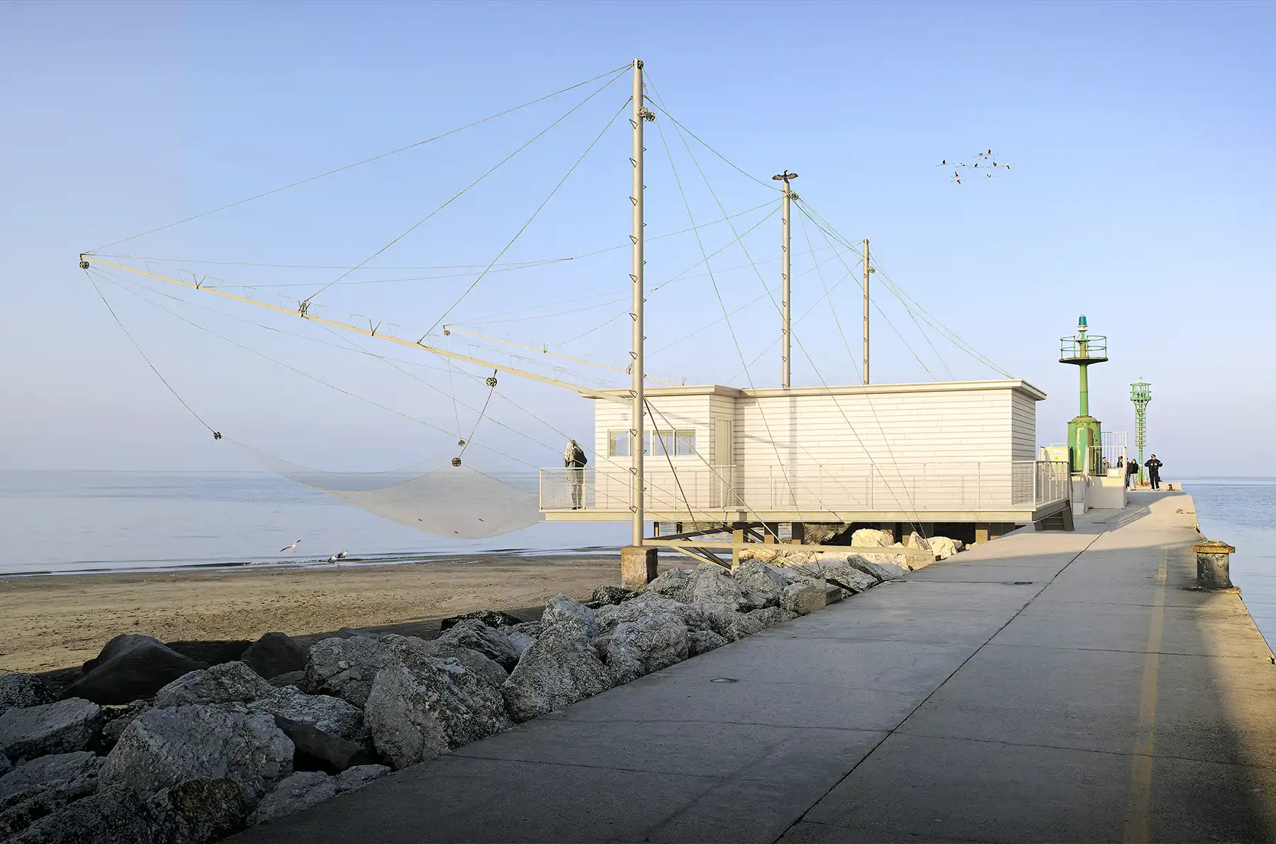 Pier leading out to the sea with a structured, modern fishing hut featuring taut cables and a large fishing net. The scene captures a serene winter sunset, an expansive view of the open water meeting the horizon, ideal for coastal architecture and peaceful seaside moments.