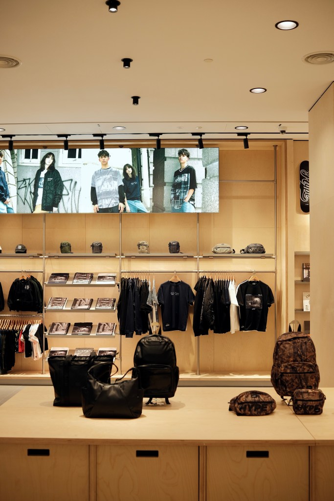 A modern clothing store interior featuring a variety of apparel hanging on racks and a display of backpacks on a wooden counter, with a large screen showing four models wearing trendy outfits in the background.