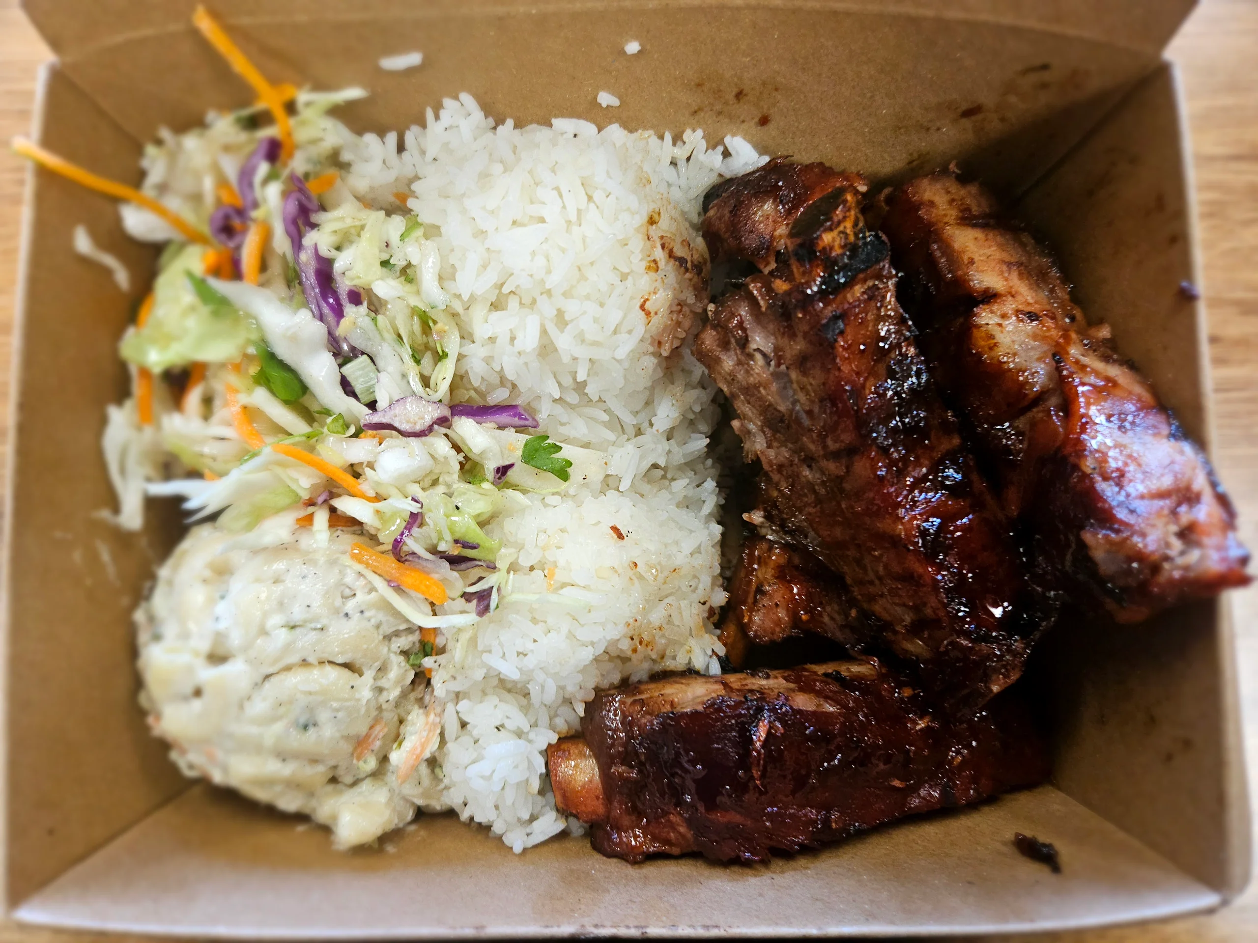 A row of tender, red-glazed Char Siu pork ribs served with two scoops of rice, macaroni salad, and a side of Asian slaw.
