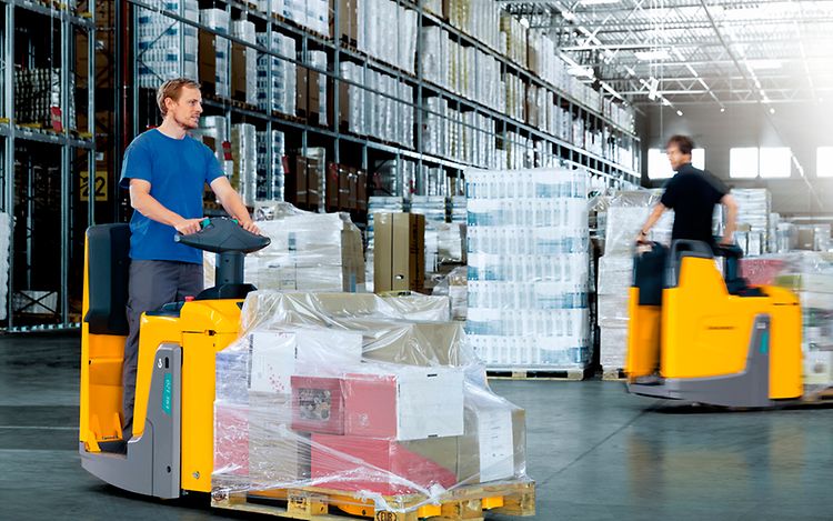 Two people in a workshop, one standing and smiling while the other leans over a car, working on it.