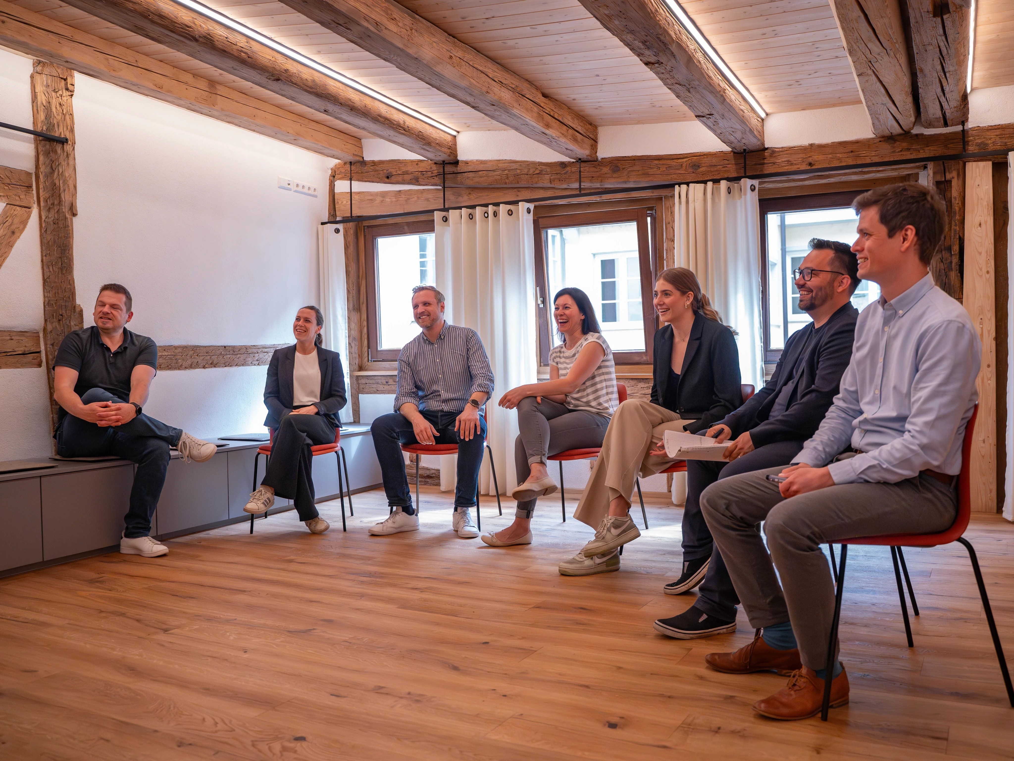 Eine Gruppe von Menschen, die in einem gut beleuchteten Raum mit Holzbalken sitzen und diskutieren oder eine Sitzung abhalten