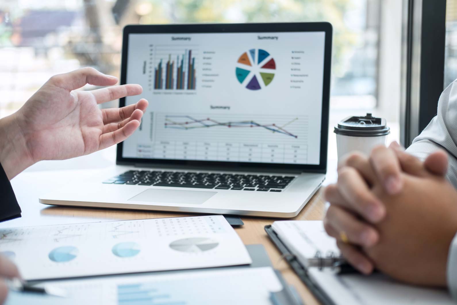 Zbliżenie na dłonie dwóch osób analizujących dane na ekranie laptopa podczas spotkania biznesowego. Na stole leżą wydrukowane raporty i kolorowe wykresy analityczne. Zdjęcie ilustruje analityczne podejście do planowania strategii dla takich projektów jak branding web design czy nowoczesne animacje motion realizowanych przez profesjonalną agencję reklamową we Wrocławiu.