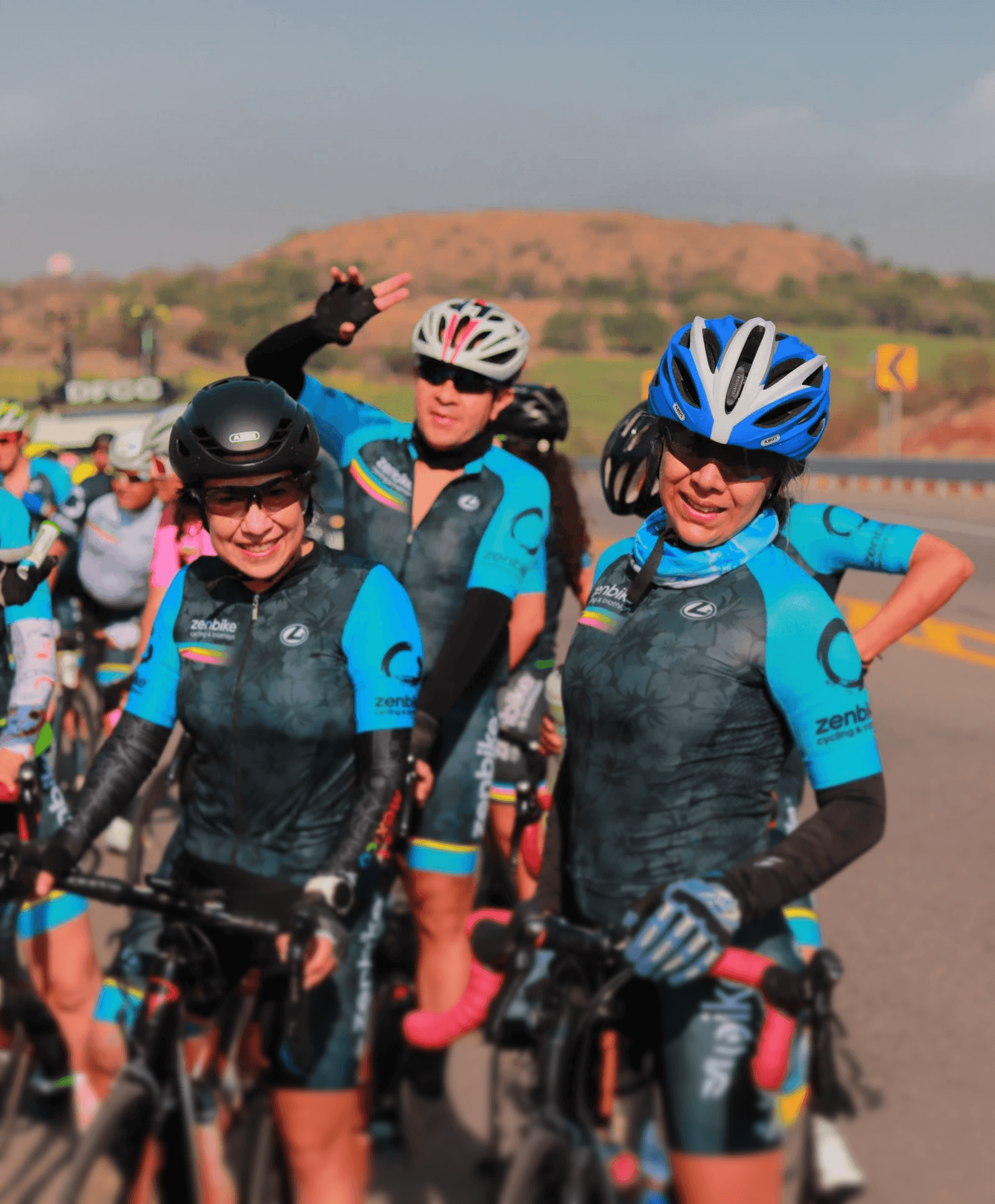 Ciclistas ZenBike con uniforme floral azul posan sonrientes durante rodada grupal en carretera panorámica.