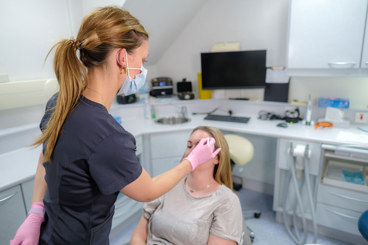 A female dental professional, wearing dark blue scrubs, a face mask, and pink gloves, uses a white marker to draw small points on the forehead of a female patient. The patient is sitting in a dental chair in a clinic setting, and the markings are being made in preparation for a facial aesthetic treatment.