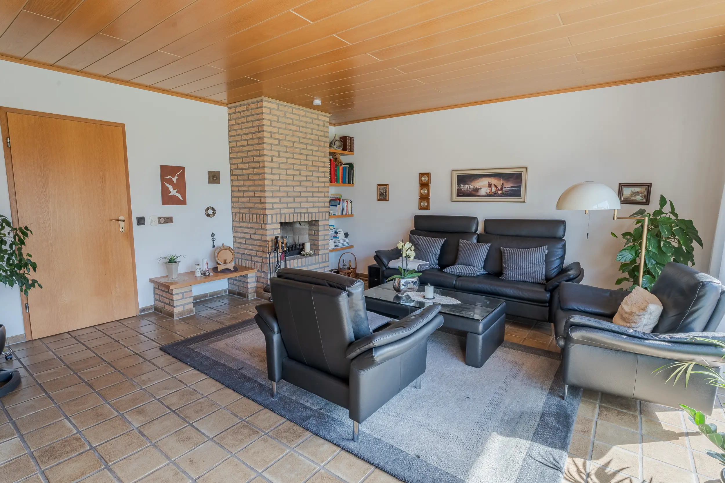 Wohnzimmer der Doppelhaushälfte in Gronau mit gemauertem Kamin, schwarzer Sitzgruppe, Holzdecke und Fliesenboden