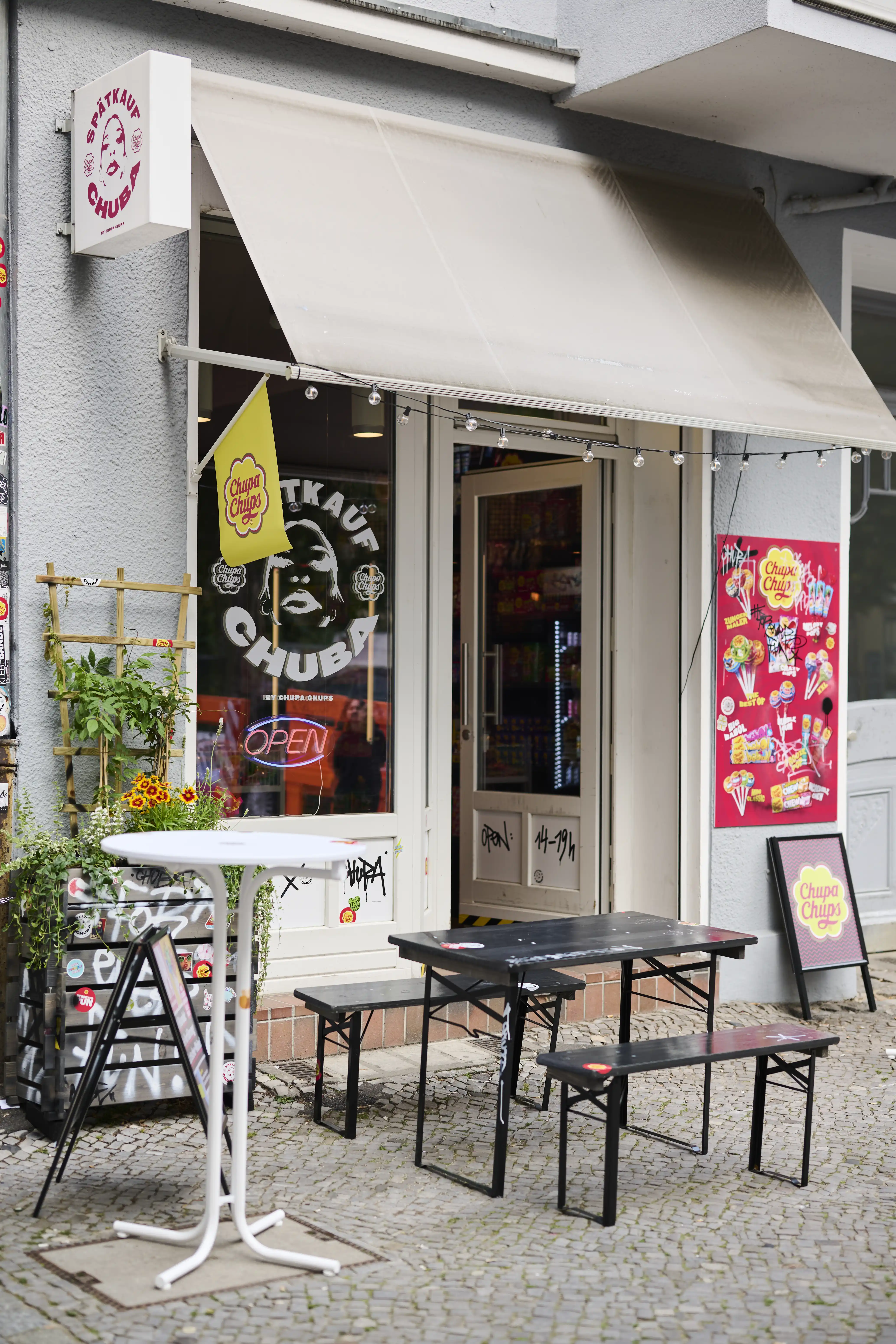 Street view of the Spätkauf Chuba pop-up kiosk by Chupa Chups with awning, beer benches and yellow brand flag in Berlin.