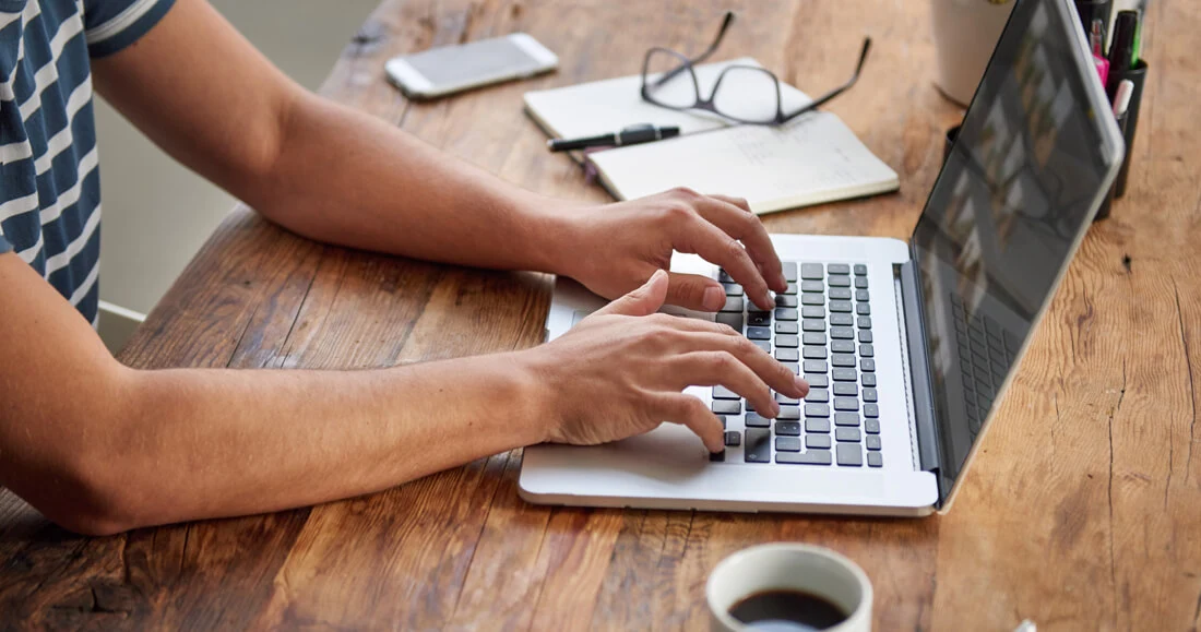 Person typing on a laptop at a wooden desk with notebook, glasses, phone, and coffee cup