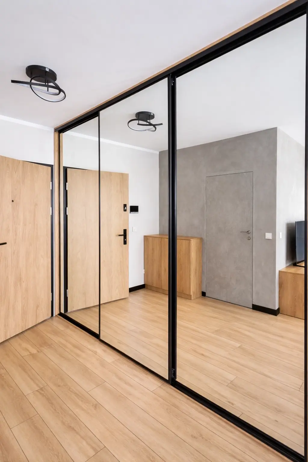 Szafa przesuwna z dwoma dużymi lustrzanymi panelami w czarnych ramach obok drzwi o wzorze drewna.