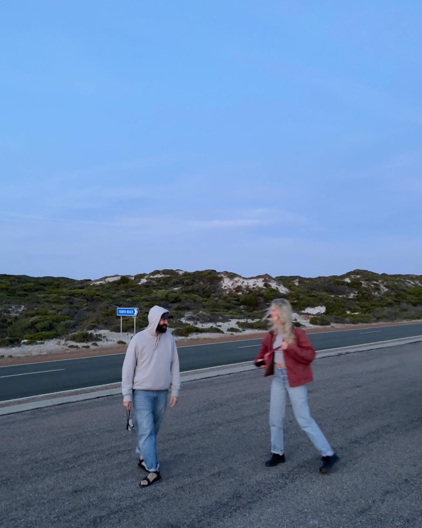 a man wearing jeans and a grey hoodie walking in the street with a blonde woman wearing jeans and a red leather jacket