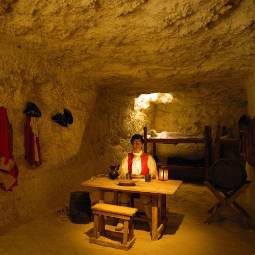 Soba nalik špilji s osobom koja sjedi za drvenim stolom. Odjeća i šeširi vise na zidu; vidljivi su krevet na kat i bačva.