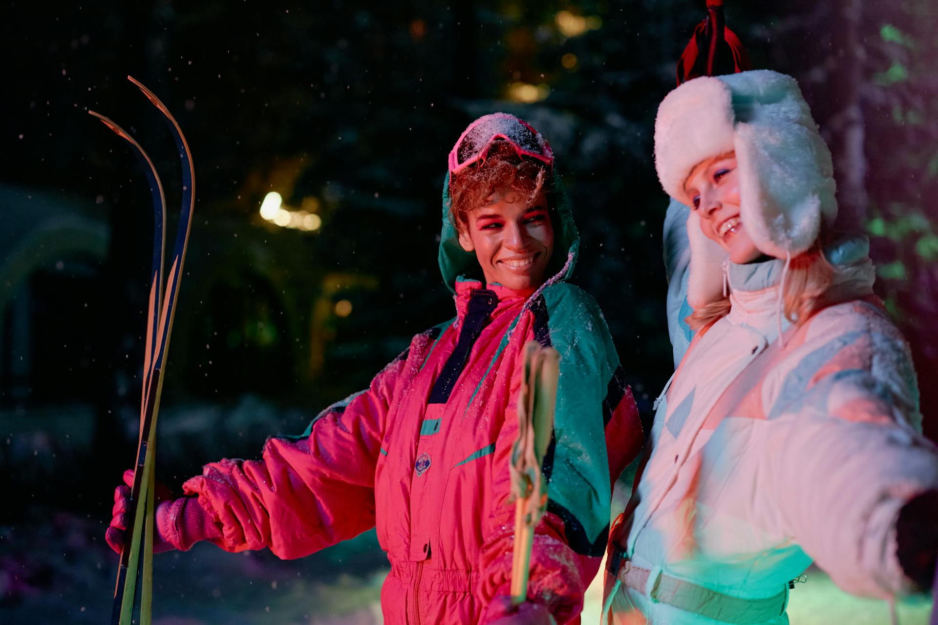 Two people in vibrant, retro-style ski suits and winter gear smile at each other while holding skis at night during a snowfall.