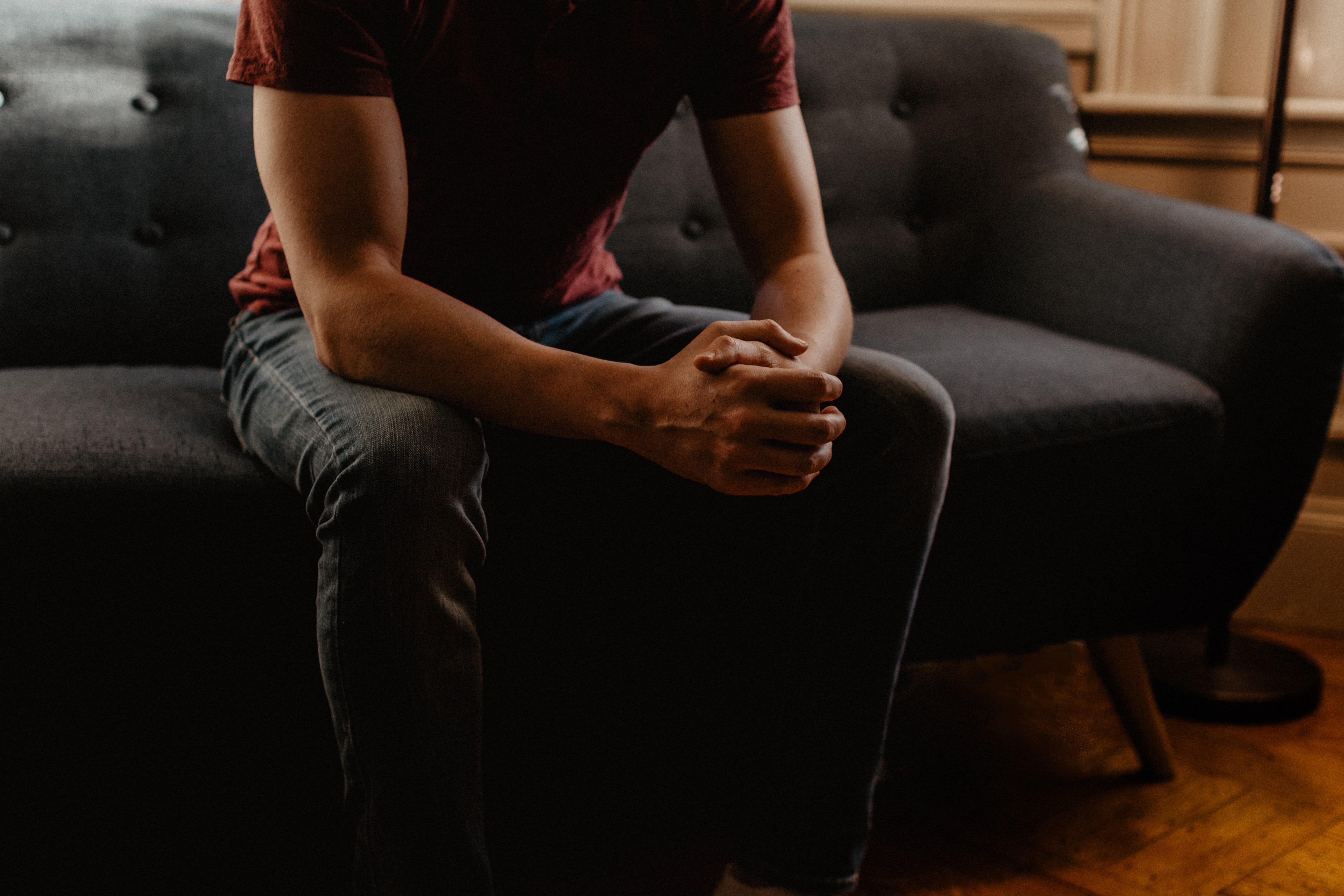 An image of a man from the neck down sat on a couch, hands clasped in front of him 