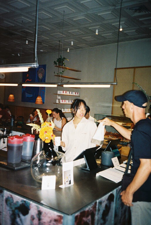 a female barista in a white button down shirt hands a coffee to a man in a black shirt and black baseball cap