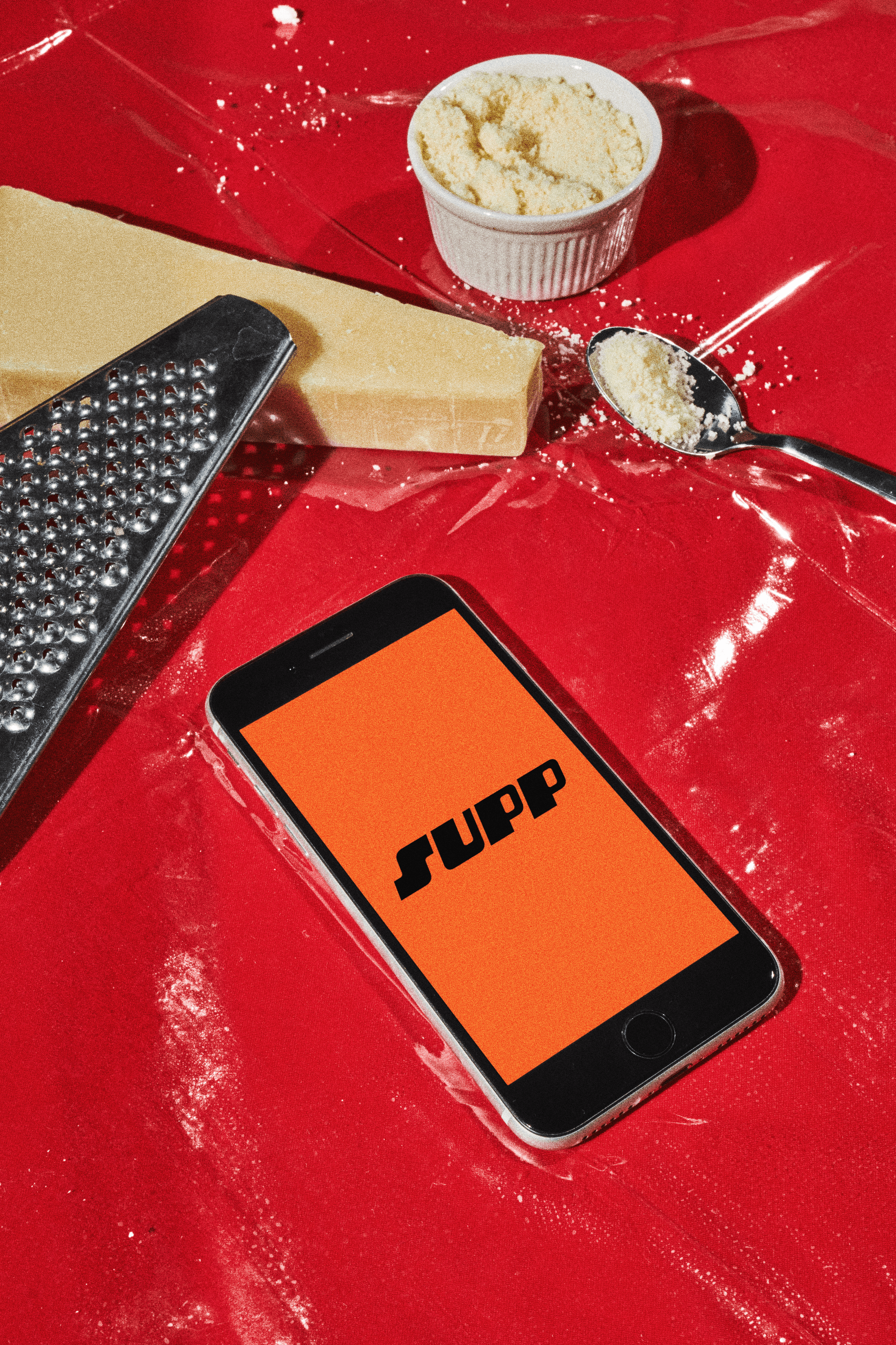 A phone displaying the Supp app logo on a red surface surrounded by a cheese grater and parmesan