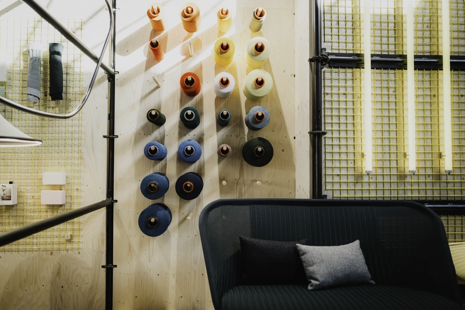 Colourful yarn cones mounted on a plywood wall above a dark knit sofa with grey cushions.