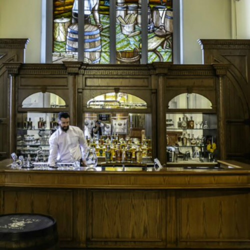 A bartender in a white shirt stands behind a wooden bar with bottles and glasses. A stained glass window is visible behind him.
