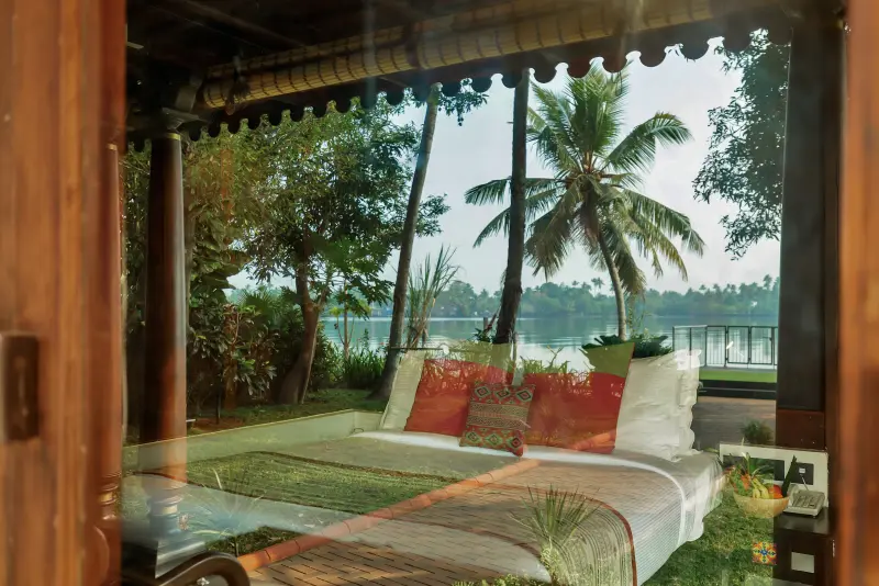 Heritage Room at Vedic Village Kodungallur with lake view through open carved wooden doors, palm trees and traditional cushions on the bed