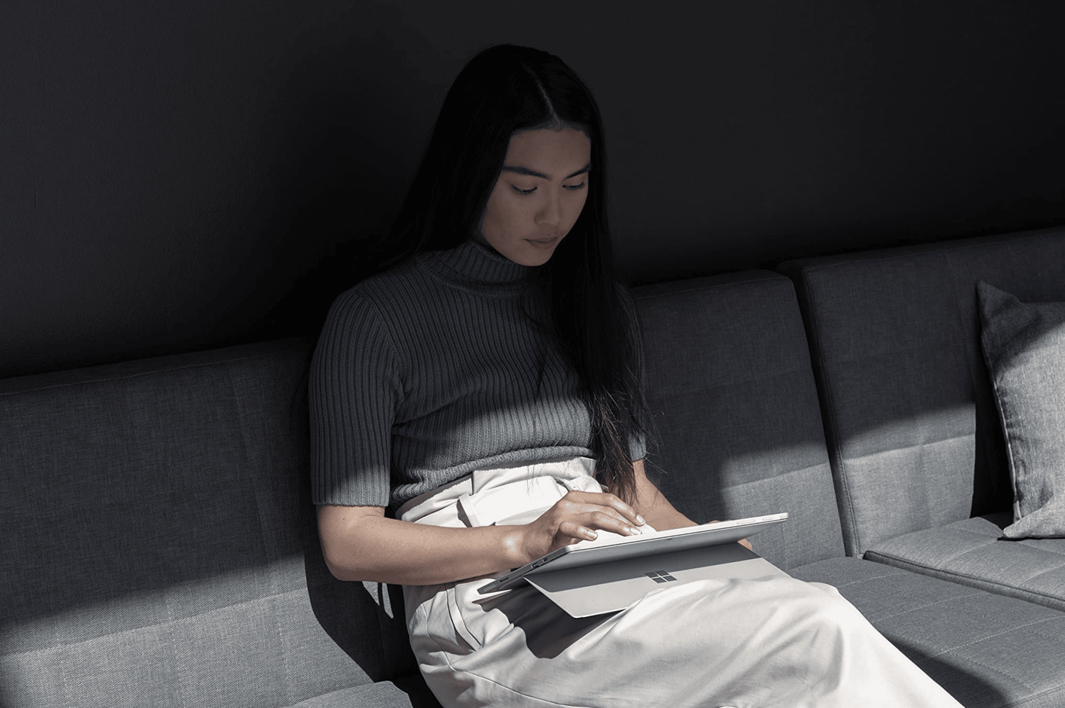 A woman using a Surface tablet while sitting on a dark grey sofa, highlighted by a dramatic beam of sunlight.