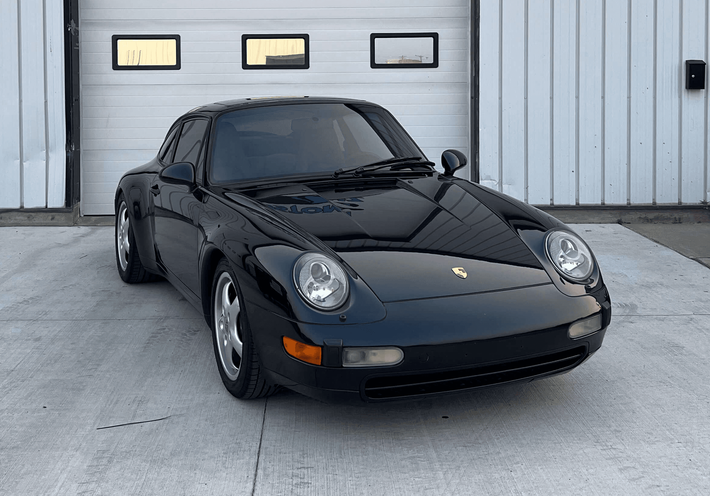 A black Porsche 911 (993 generation) sports car is parked on a concrete driveway in front of a white industrial building.