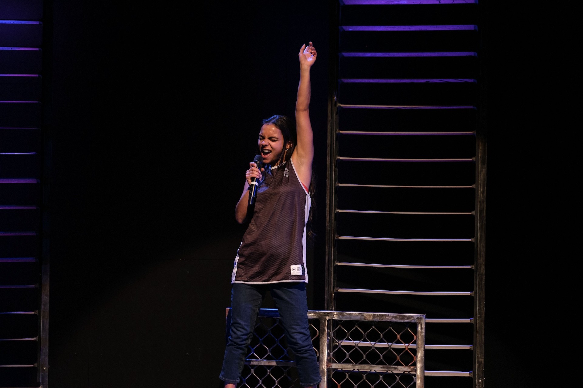 A performer stands confidently with one arm raised, singing into a microphone. She wears a basketball singlet and jeans, framed by tall metal scaffolding under purple lighting.