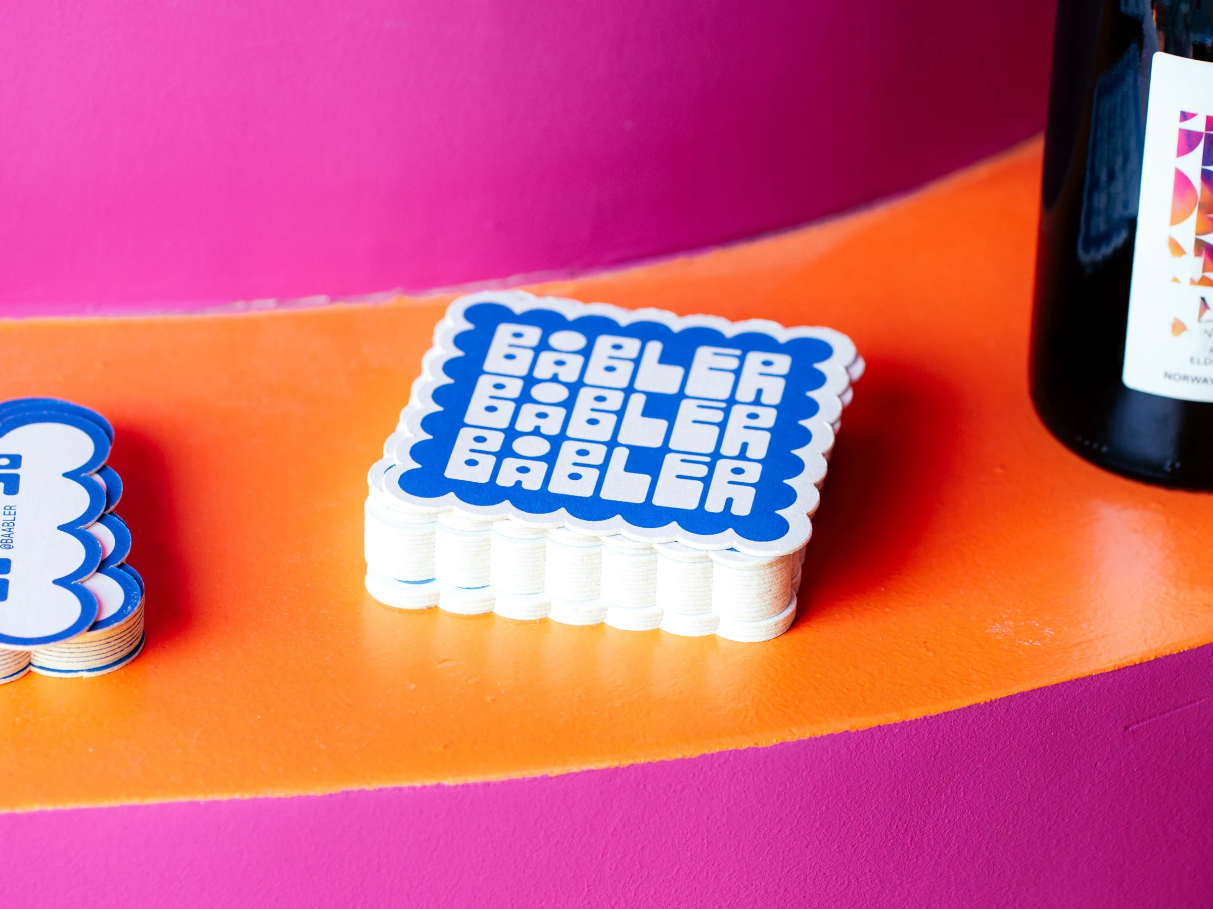 Coaster designs for Båbler wine bar laying in a stack on a orange and pink table.