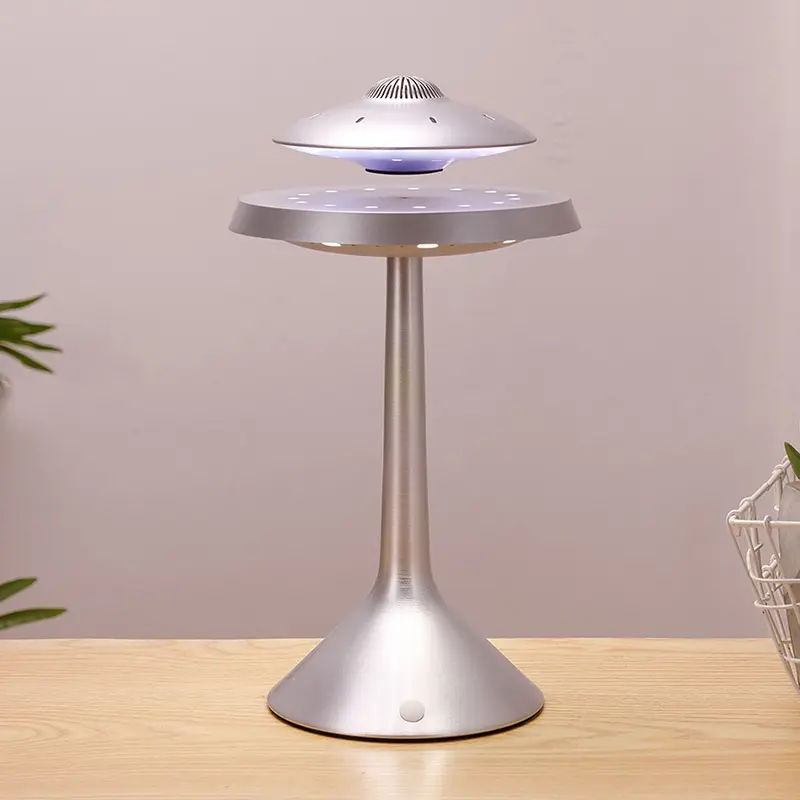 A sleek, modern LED lamp with a hovering UFO-shaped light hovering above its metallic base. The design creates a levitating illusion. The lamp is placed on a wooden table, with a plant in the background.