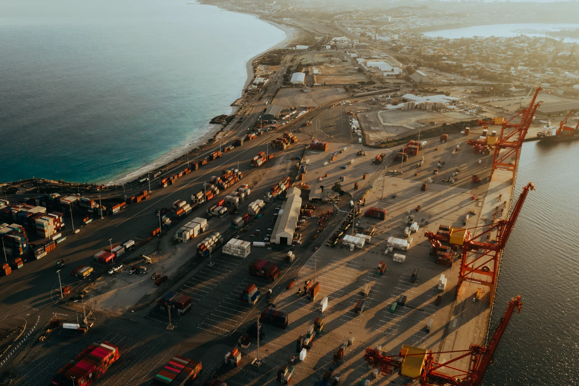 Aerial of a port yard at low sun, trucks and containers casting long shadows beside a crane