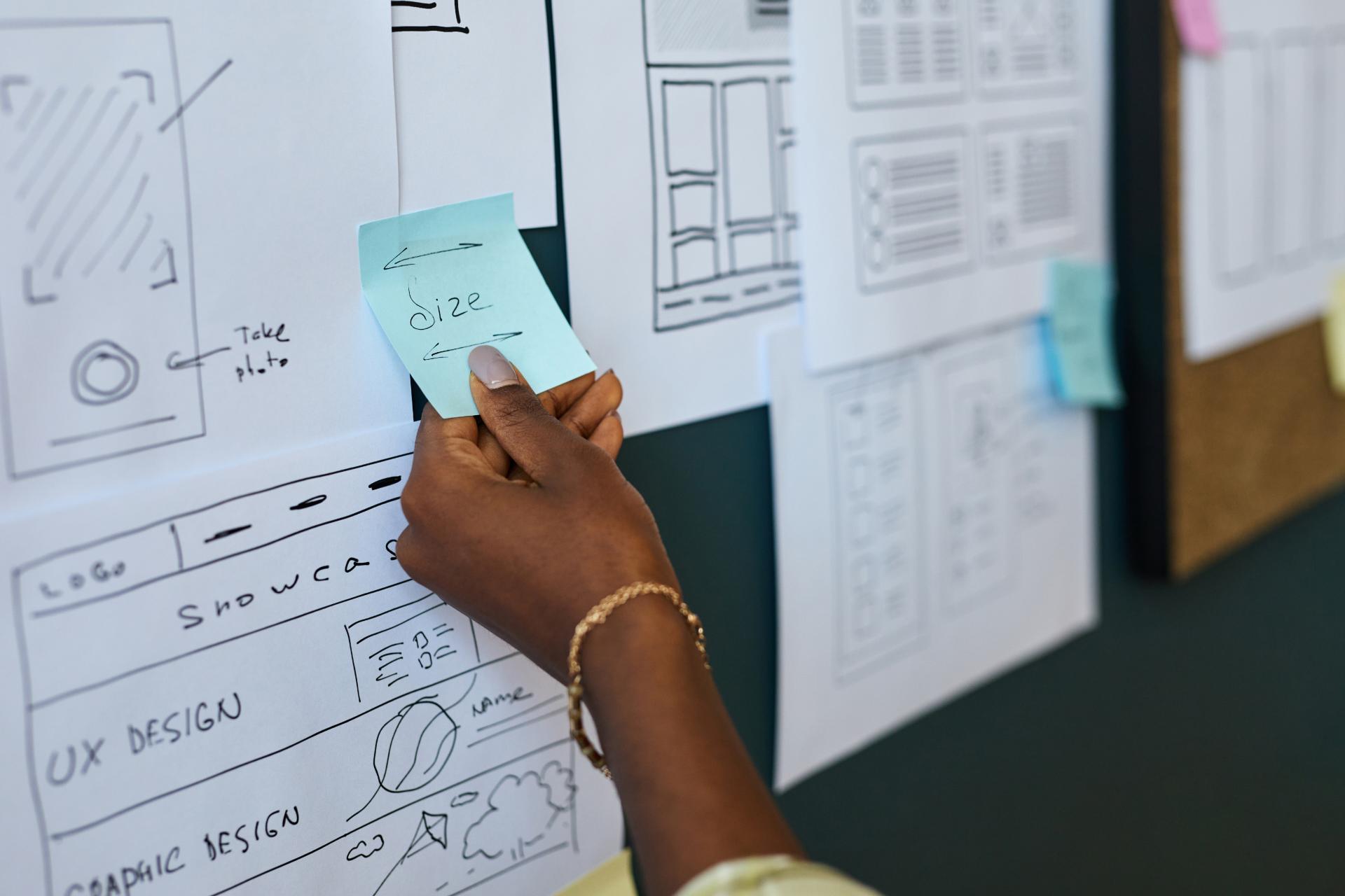 Black woman hand holding sticky note with word size written on it placing note on wall covered with wireframe sketches and user interface design layouts during creative planning session