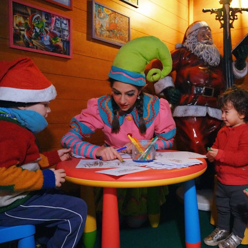 Eine als Elf verkleidete Person hilft zwei Kindern beim Ausmalen an einem Tisch, während im Hintergrund eine Weihnachtsmannstatue steht.