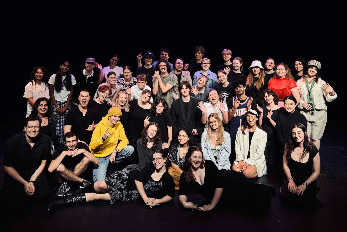 A large group of young people and staff members from ATYP gather onstage for a group photo, smiling and posing together under theatrical lighting. The group fills the lower half of the frame, with a dark background above and warm stage lights glowing overhead.