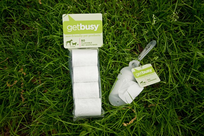 A package of Getbusy pet waste bags lies on a patch of green grass. The package contains four rolls of pet waste and is labeled “eco-friendly pet products” and “80 flushable pet waste bags.” A small tag attached to the package reads “getbusy” with “bag dispenser” written below. The package is partially open, revealing the rolls inside.