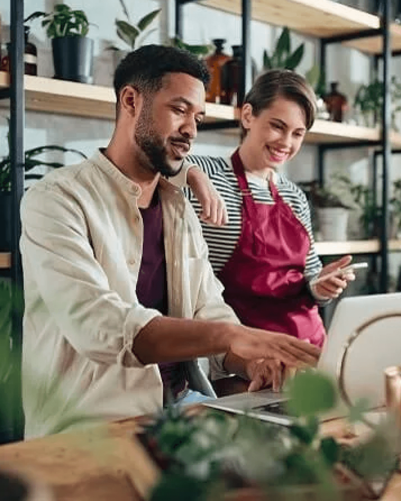 A professional man and woman reviewing a laptop screen together in a workspace with a background of potted plants on a rack