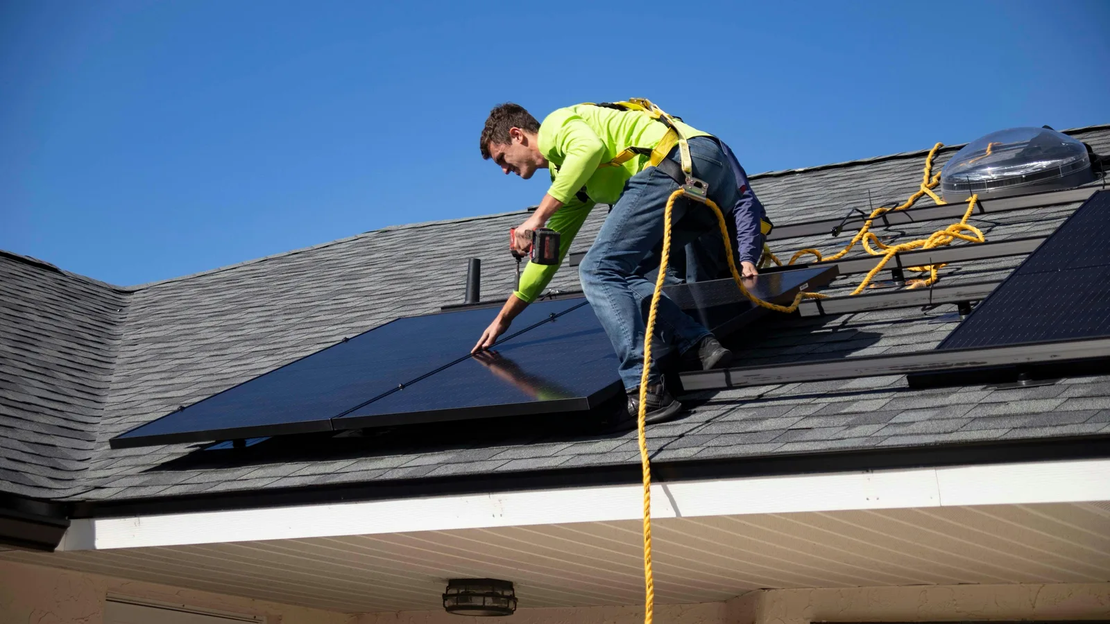 Technician in high-visibility vest installing solar panel on roof with drill