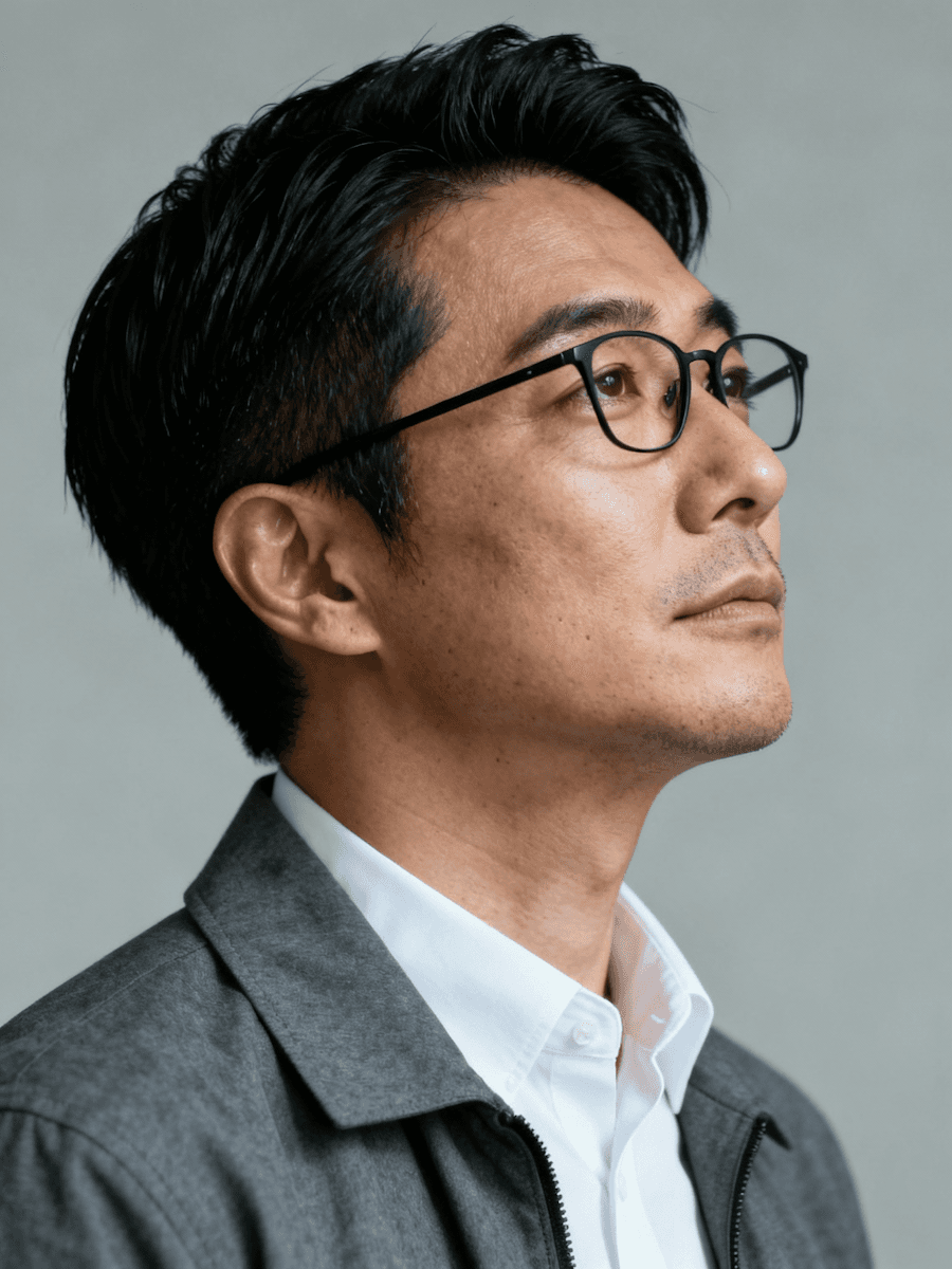 A man with glasses and a thoughtful expression, wearing a white shirt and gray jacket, captured in a neutral background.