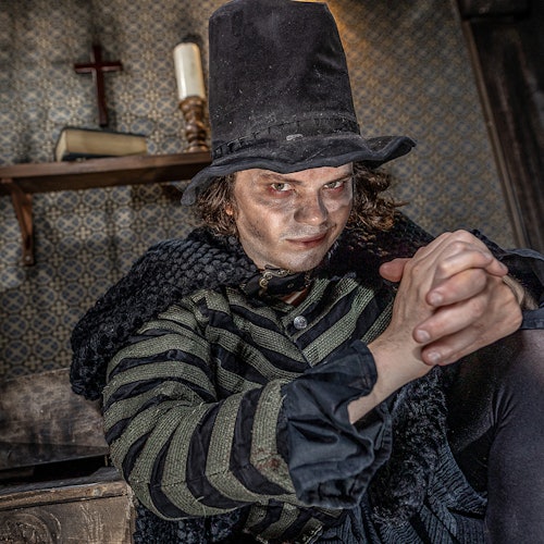 一个穿着条纹服装和高帽子的人专注地凝视着,坐在一个昏暗的房间里,背后的架子上放着一个十字架和蜡烛。