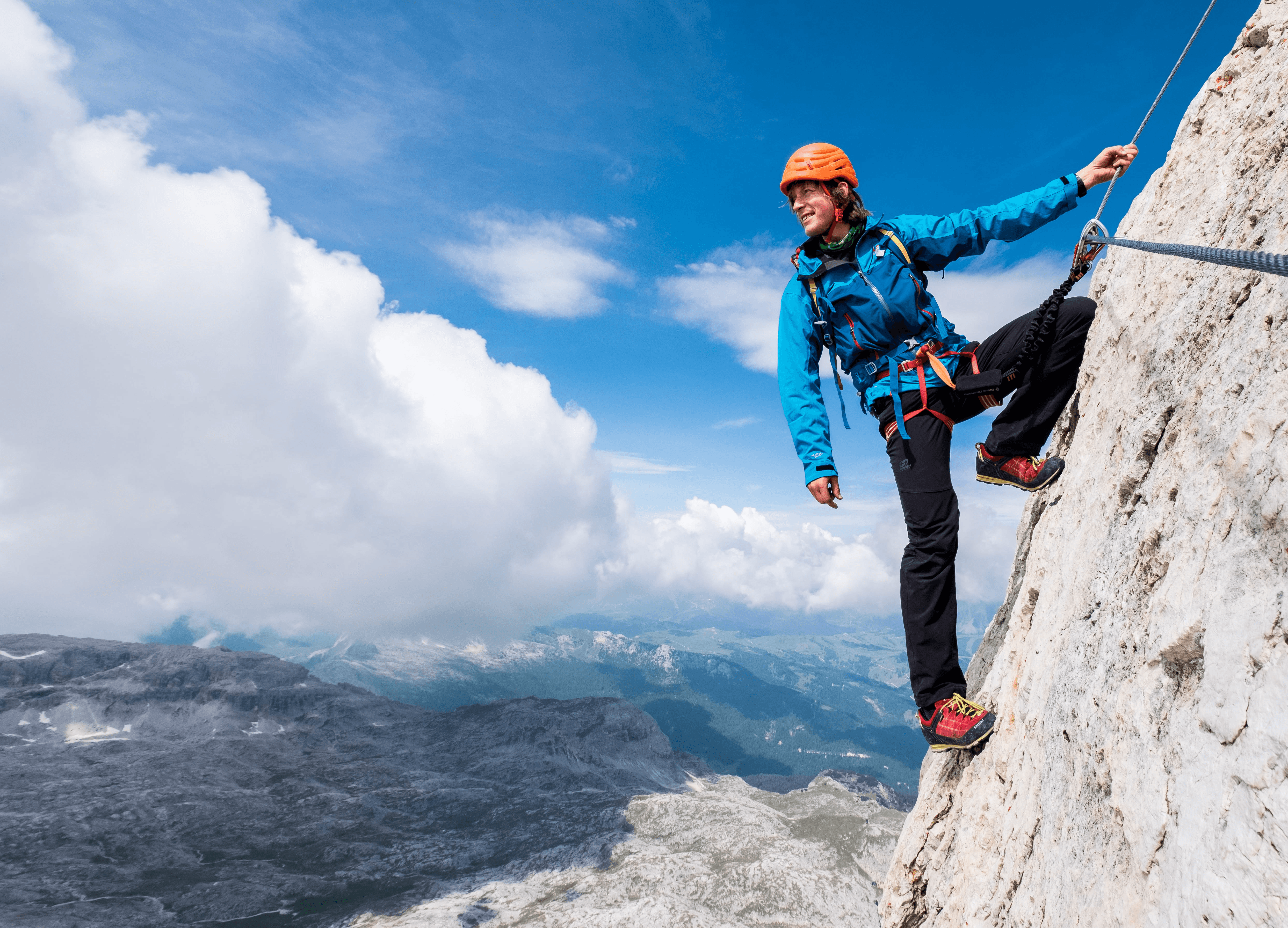 A climber wearing a blue jacket and orange helmet scaling a steep rock face with a breathtaking mountain view in the background.